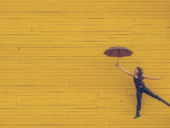 Woman jumping with umbrella in front of yellow wall.